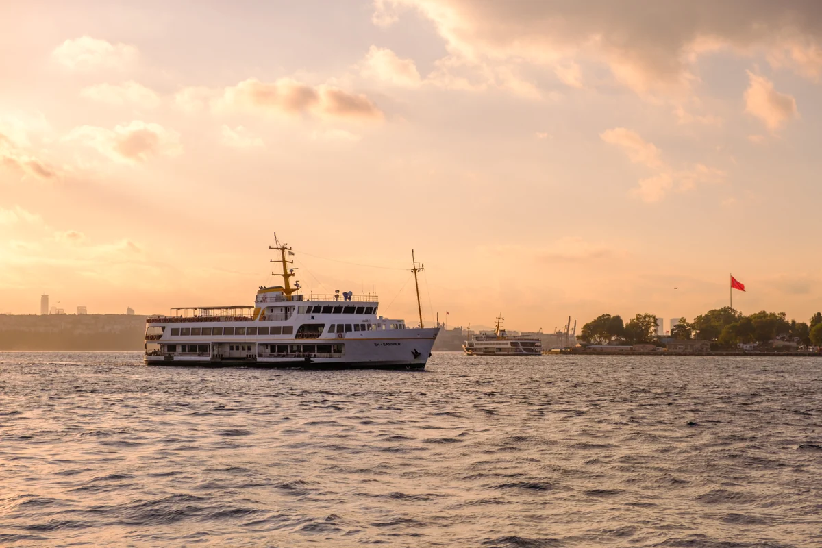 A traditional white passenger ferry navigates the Bosphorus strait in Istanbul during a hazy sunset, with the city skyline and minarets visible on the distant shore.
