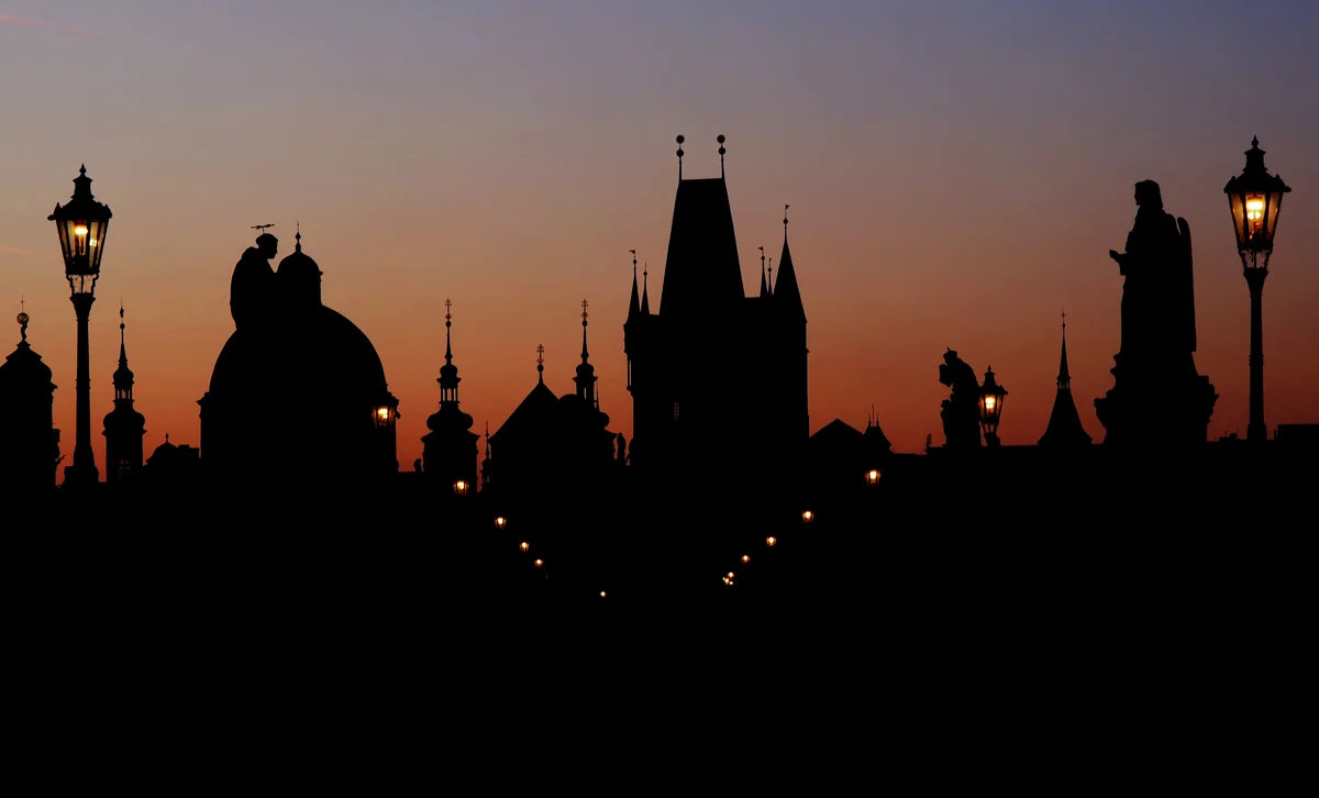 Silhouettes of Baroque statues line the Charles Bridge in Prague against a glowing orange and pink dawn sky, with the Old Town Bridge Tower and church spires in the distance.